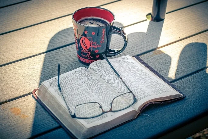 caneca preta de café com detalhes em vermelho ao lado de livro aberto com óculos em cima