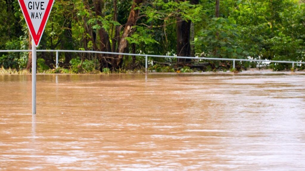 Rua alagada com uma placa vermelha parcialmente submersa e árvores ao fundo