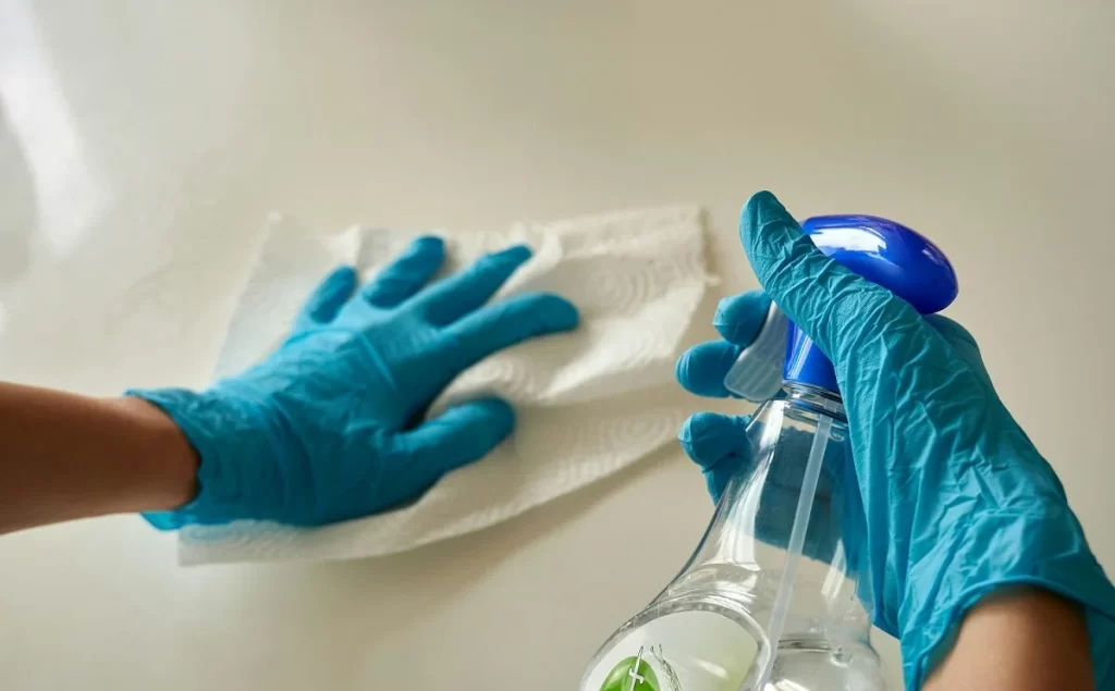 mãos com luvas azuis com um frasco de produto de limpeza transparente passando um papel toalha em uma superfície branca