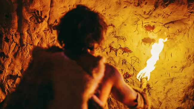Home das cavernas segurando uma tocha em frente á uma rocha com pintura rupestre