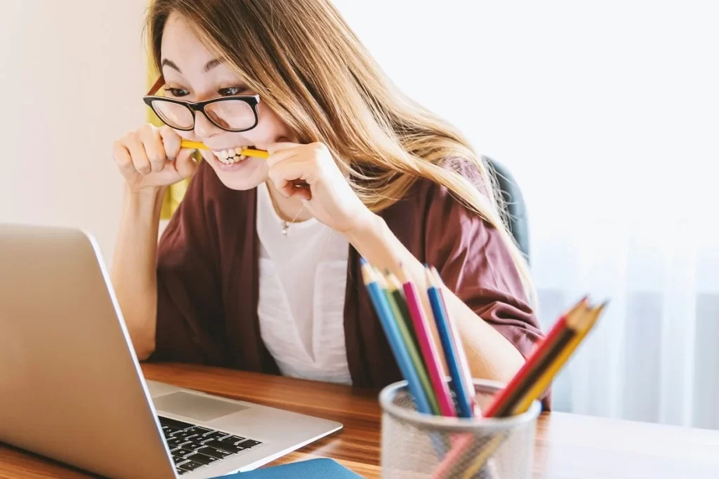 mulher de óculos mordendo um lápis amarelo com expressão de nervoso olhando para um laptop