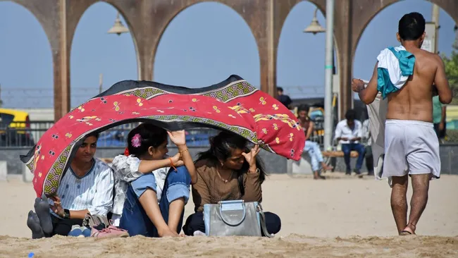 Três mulheres se protegendo do sol escaldante com um pano vermelho na areia em Mumbai, na Índia.