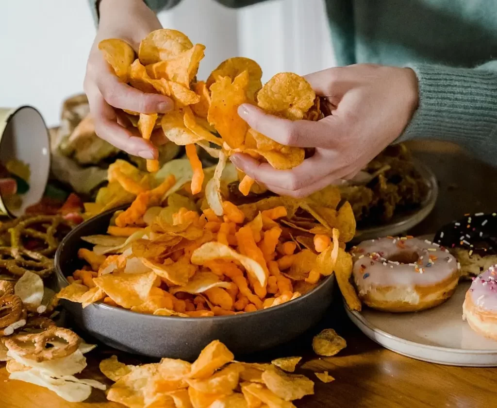 mesa cheia de junk food com uma pessoa enchendo a mão de chips
